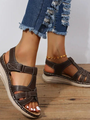 Bruine leren sandalen voor dames met klittenbandsluiting, gedragen met gescheurde jeans. Comfortabele zomerschoenen op houten vloer.