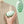 Hand houdt groene draagbare massageapparaat vast op beige trui; ergonomisch ontwerp, stressverlichting, spierontspanning, wellness gadget.
