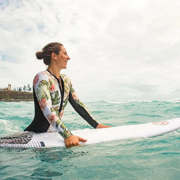 Vrouw in bloemen wetsuit op surfplank in oceaan, lachend, met golven en kustlijn op achtergrond. Surfen, watersport, strandvakantie.