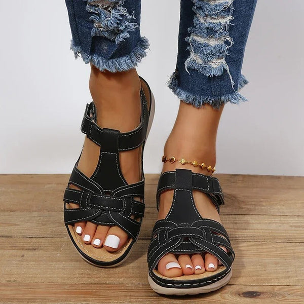 Zwarte leren sandalen voor dames met open teen en verstelbare bandjes, gedragen met gescheurde jeans. Comfortabele zomerschoenen op houten vloer.