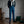 Man in stijlvolle blauwe broek en geruite blouse met witte sjaal, staand in elegante kamer met houten vloer en decoratieve elementen.