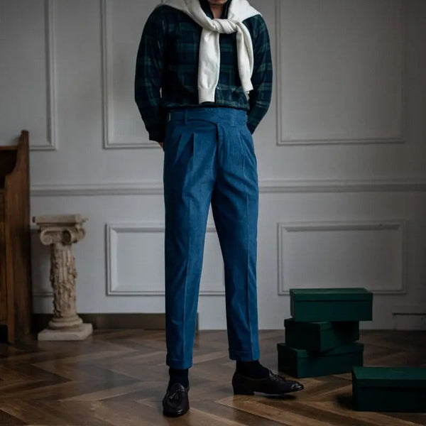 Man in stijlvolle blauwe broek en geruite blouse met witte sjaal, staand in elegante kamer met houten vloer en decoratieve elementen.