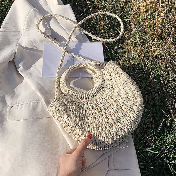 Rieten handtas met ronde handvatten op beige jas, liggend op gras. Perfect voor zomerse outfits en stranduitjes. Trendy en duurzaam design.