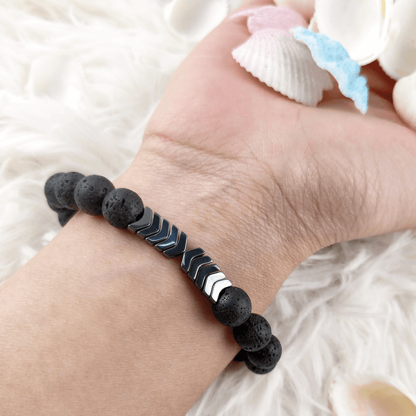 Zwart lavasteen armband met metalen pijlen, olie diffuser functie, hand op witte achtergrond, ideaal voor aromatherapie en stijlvol accessoire.