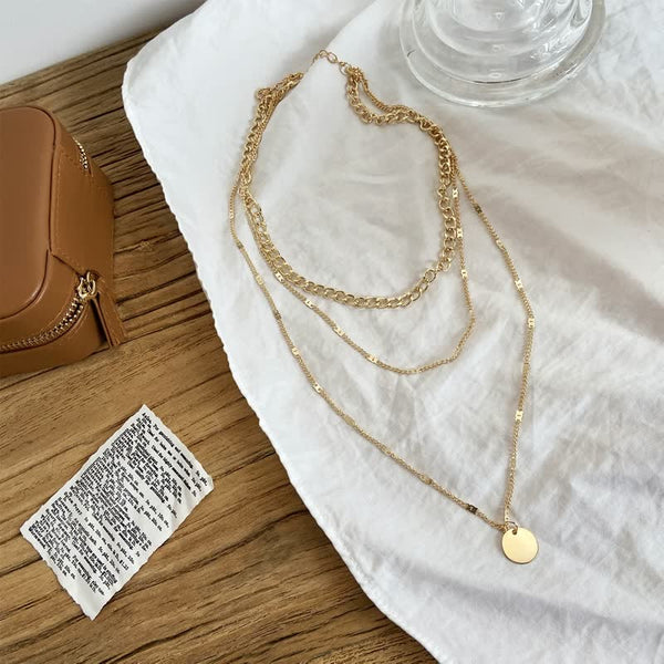 Gouden ketting met meerdere lagen en ronde hanger op witte stof, naast bruine tas en tekstbriefje. Perfecte accessoire voor modieuze stijl.