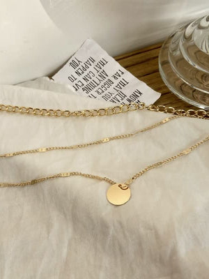 Gouden ketting met ronde hanger op witte achtergrond, naast glazen vaas en papieren label. Elegante sieraden, minimalistisch design, modetrend.