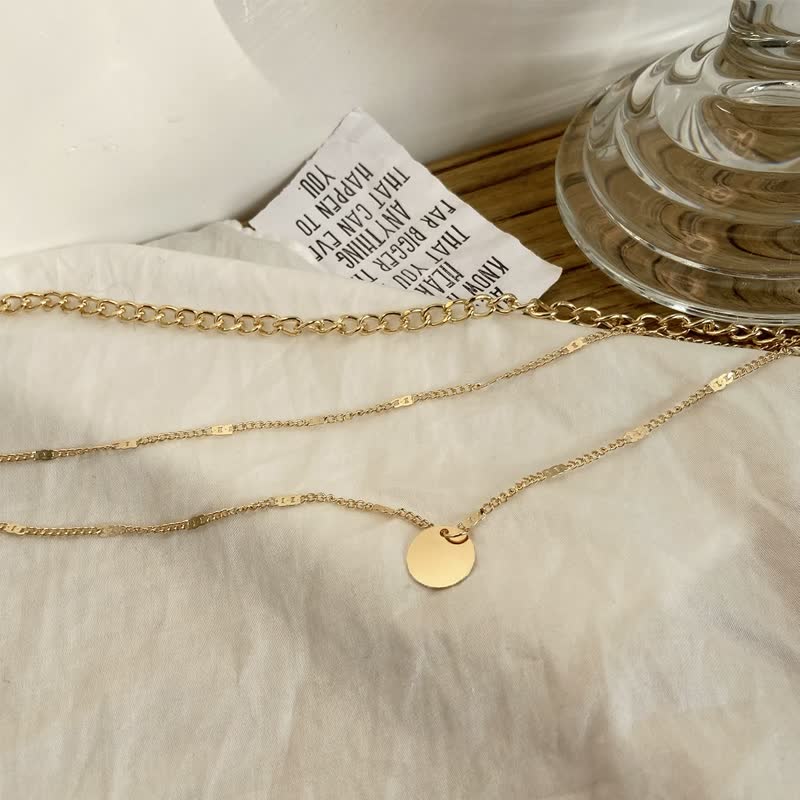 Gouden ketting met ronde hanger op witte achtergrond, naast glazen vaas en papieren label. Elegante sieraden, minimalistisch design, modetrend.