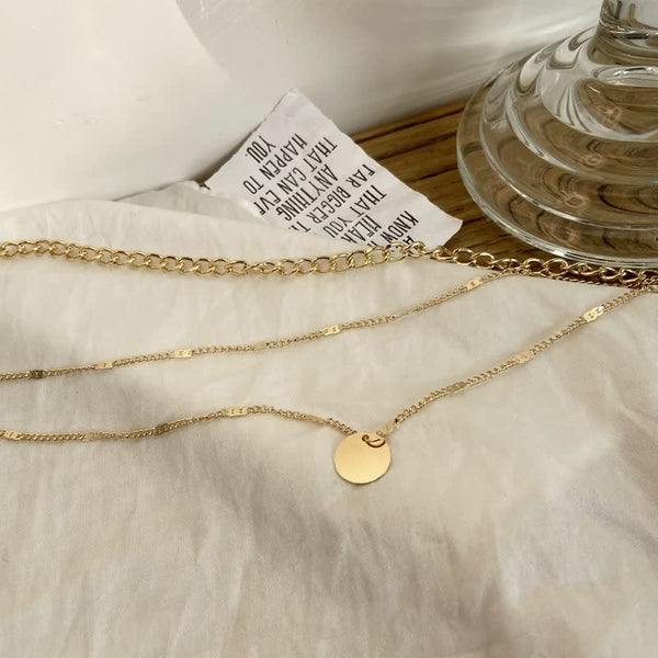 Gouden ketting met ronde hanger op witte achtergrond, naast glazen vaas en papieren label. Elegante sieraden, minimalistisch design, modetrend.