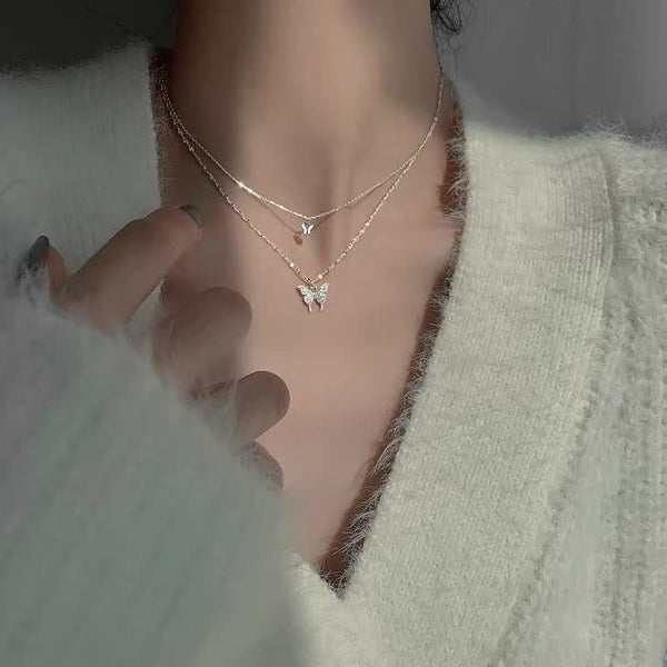 Twee delicate vlinderkettingen op een hals, gedragen met een zachte, witte trui. Perfecte sieraden voor een elegante en modieuze look.