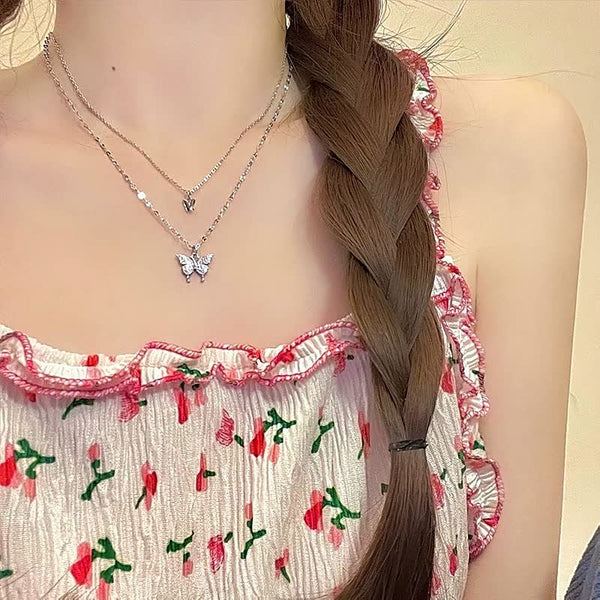 Vrouw met vlecht draagt zilveren kettingen met vlinder- en hartjeshanger, gekleed in bloemenjurk. Modeaccessoires, sieraden, zomerstijl.