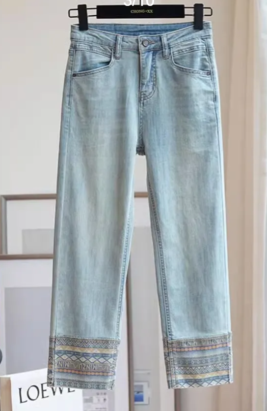 Lichtblauwe spijkerbroek met etnische borduursels op de zoom, hangend aan een hanger. Trendy damesmode, casual denim stijl.