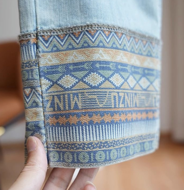 Hand houdt denim broekspijp met kleurrijk etnisch patroon vast, inclusief geometrische vormen en tribal motieven. Perfect voor boho mode en casual stijl.