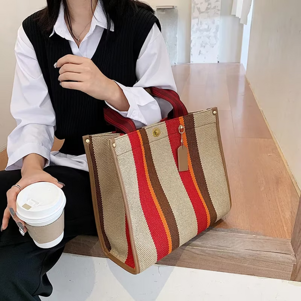 Vrouw met gestreepte canvas tas in rood, oranje en beige tinten, zittend met koffiebeker. Modieuze handtas, casual outfit, binnenomgeving.