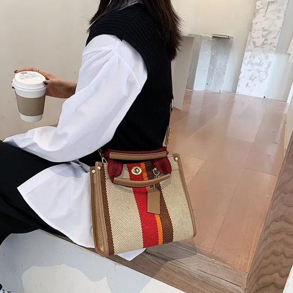 Vrouw met stijlvolle, gestreepte handtas in rood, beige en bruin, zittend op houten trap met koffiebeker. Modieuze accessoires, casual outfit.