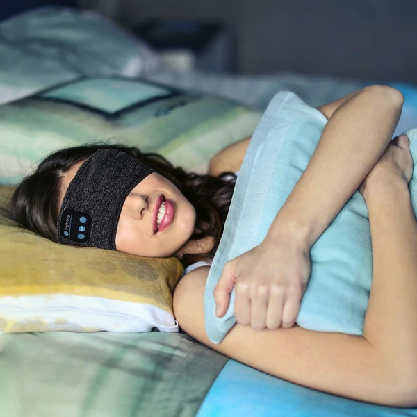 Vrouw draagt zwarte slaapband op bed, ideaal voor slaap en sport. Comfortabele hoofdband met draadloze technologie. Perfect voor ontspanning.
