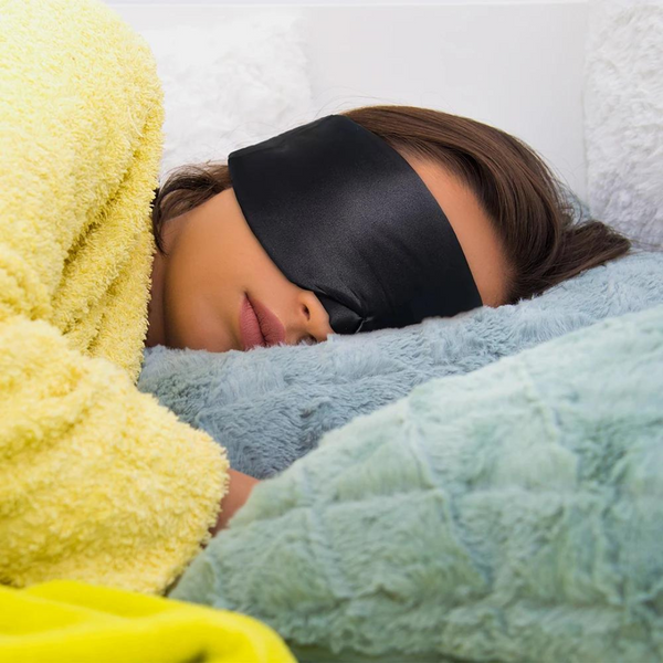 Vrouw slaapt met zwarte slaapmasker, omringd door zachte kussens en gele deken. Comfortabel slapen, slaapaccessoires, rustgevende nachtrust.