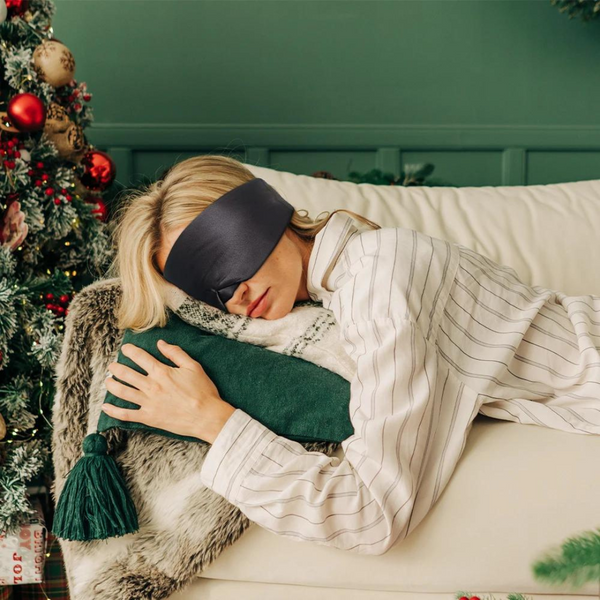 Vrouw met slaapmasker slaapt op bank, omringd door kerstdecoraties. Comfortabele pyjama, groene kussen en feestelijke sfeer. Perfect voor ontspanning.