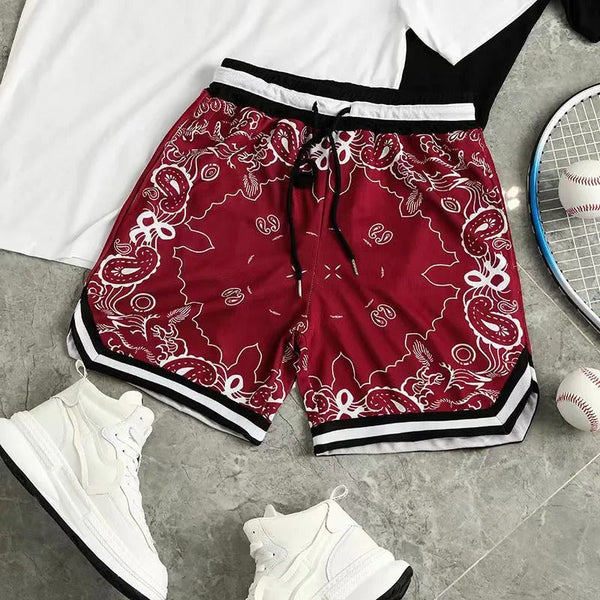 Rode bandana-print sportshorts met zwarte en witte strepen, naast witte sneakers en een tennisracket op een grijze vloer.