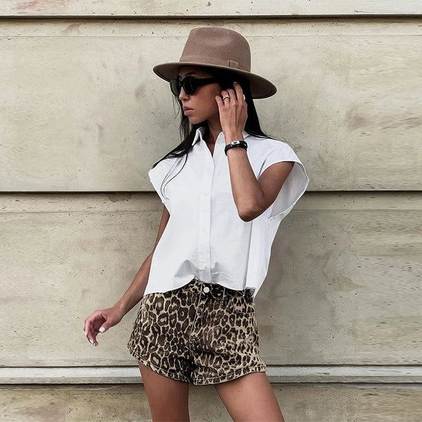 Vrouw in witte blouse en luipaardprint shorts, draagt zonnebril en hoed, poseert tegen beige muur. Zomermode, casual stijl, trendy outfit.