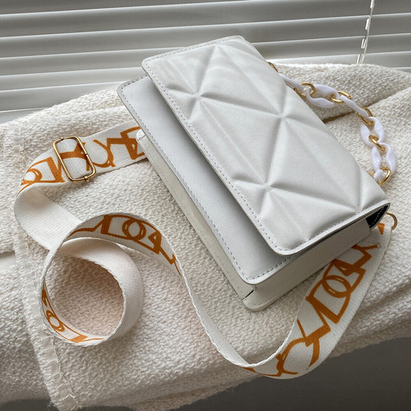 Witte gewatteerde schoudertas met gouden kettingriem en oranje logo, liggend op een crèmekleurige stof. Modieuze damesaccessoire.