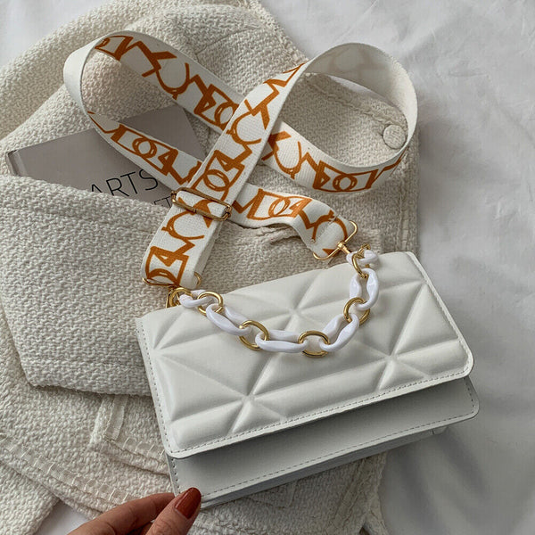 Witte leren handtas met gouden ketting en verstelbare schouderriem met oranje patroon, liggend op een crèmekleurige stoffen achtergrond.