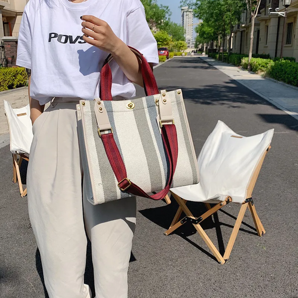 Vrouw met gestreepte canvas tas met rode schouderband op zonnige straat. Modieuze handtas, casual outfit, buiten, zomerse stijl.