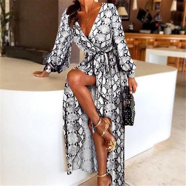 Vrouw in stijlvolle wikkeljurk met slangenprint, V-hals en split, zittend op een moderne bank. Perfecte zomerjurk voor dames.