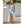 Elegante mouwloze blauwe jurk met asymmetrische zoom, gedragen door model naast witte vaas en bloemen. Perfect voor zomerse gelegenheden.